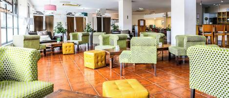 A spacious lobby with green patterned chairs, a yellow ottoman, and a tiled floor.