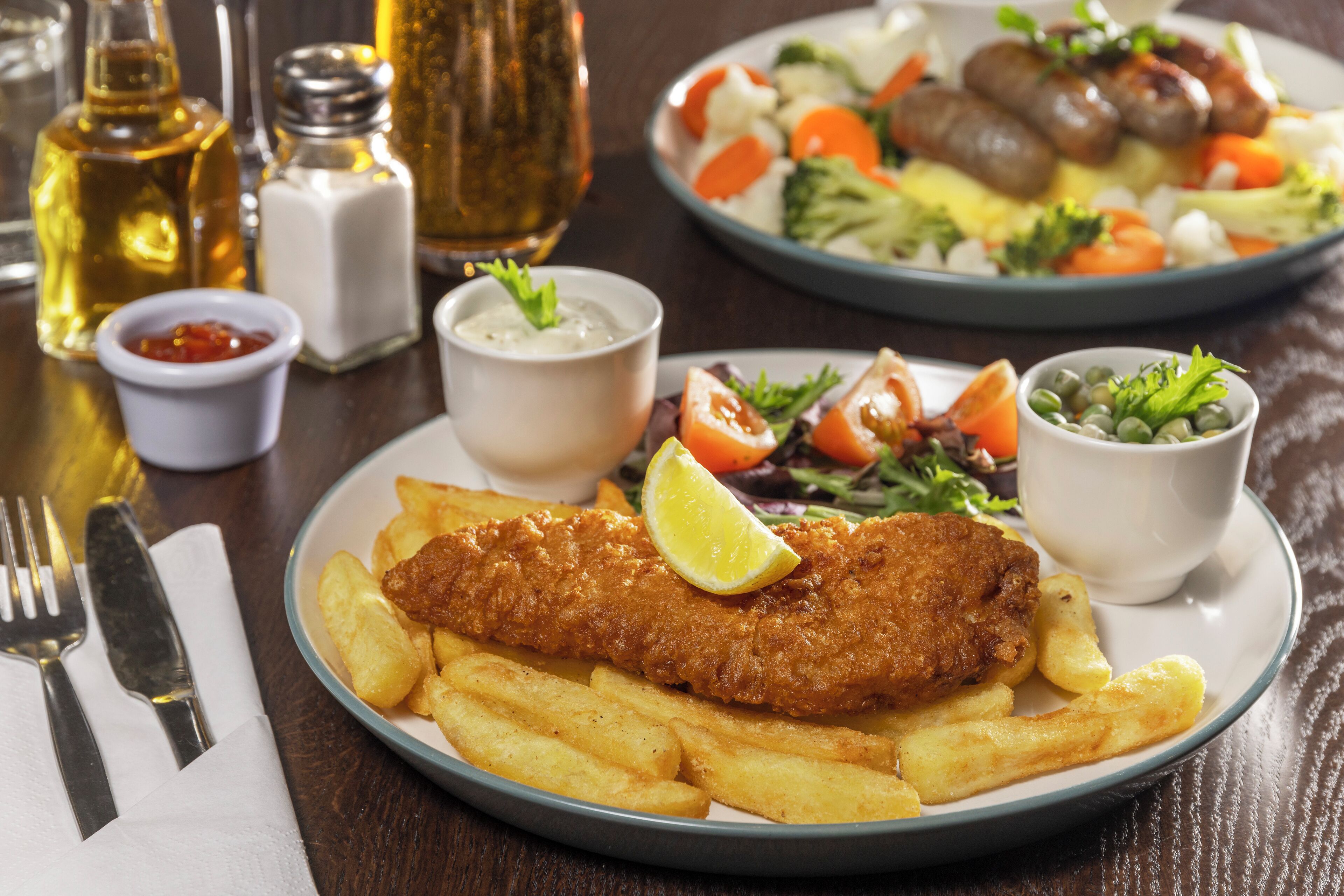 A plate of fish and chips with fries, a salad, and condiments on a table.