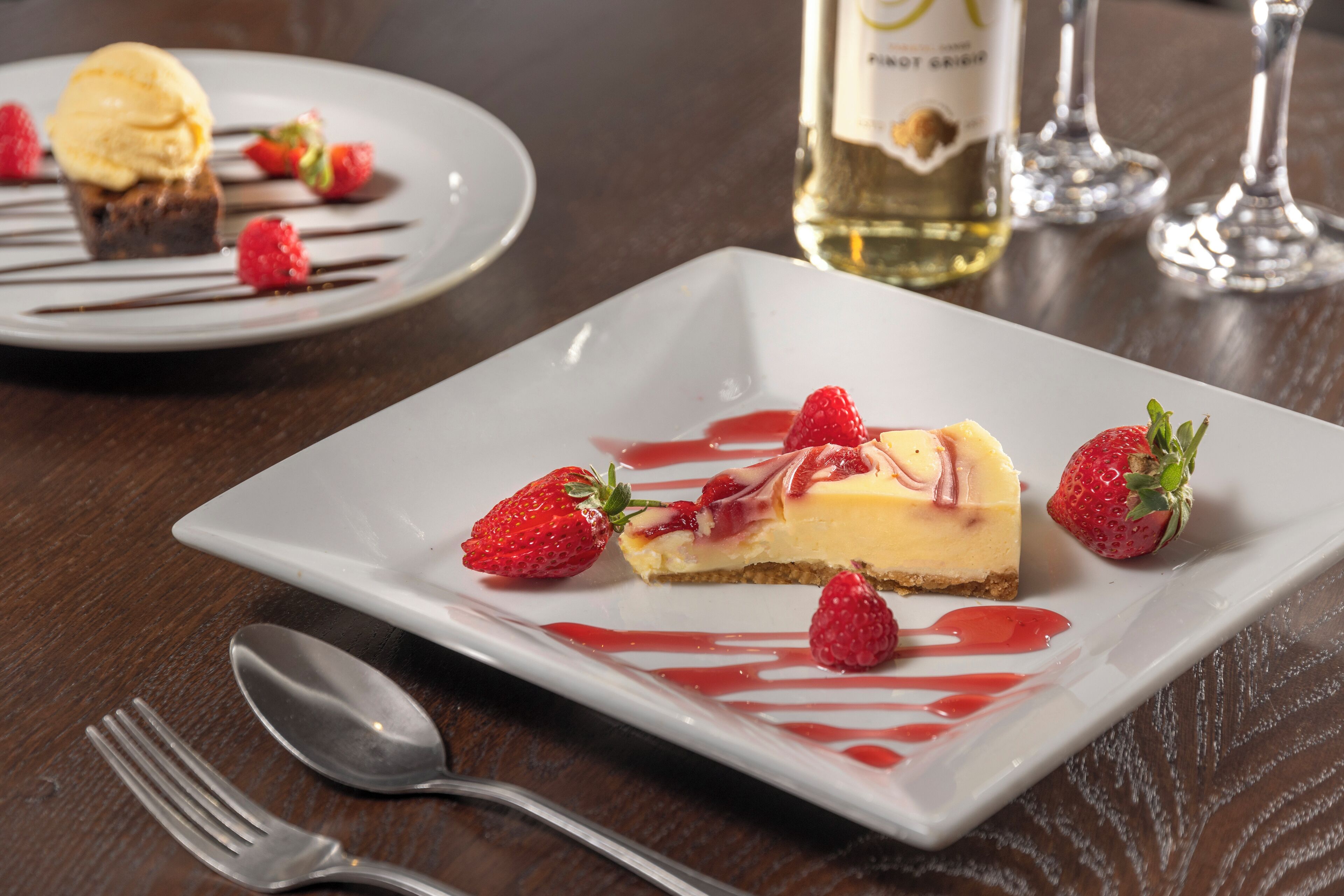 A dessert plate with a slice of cheesecake, strawberries, and a drizzle of sauce, accompanied by a fork and spoon on a wooden table.