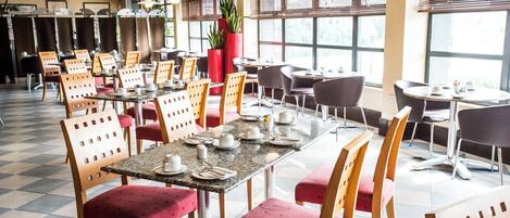 Un comedor amplio con varias mesas y sillas, ventanas grandes y plantas en macetas.