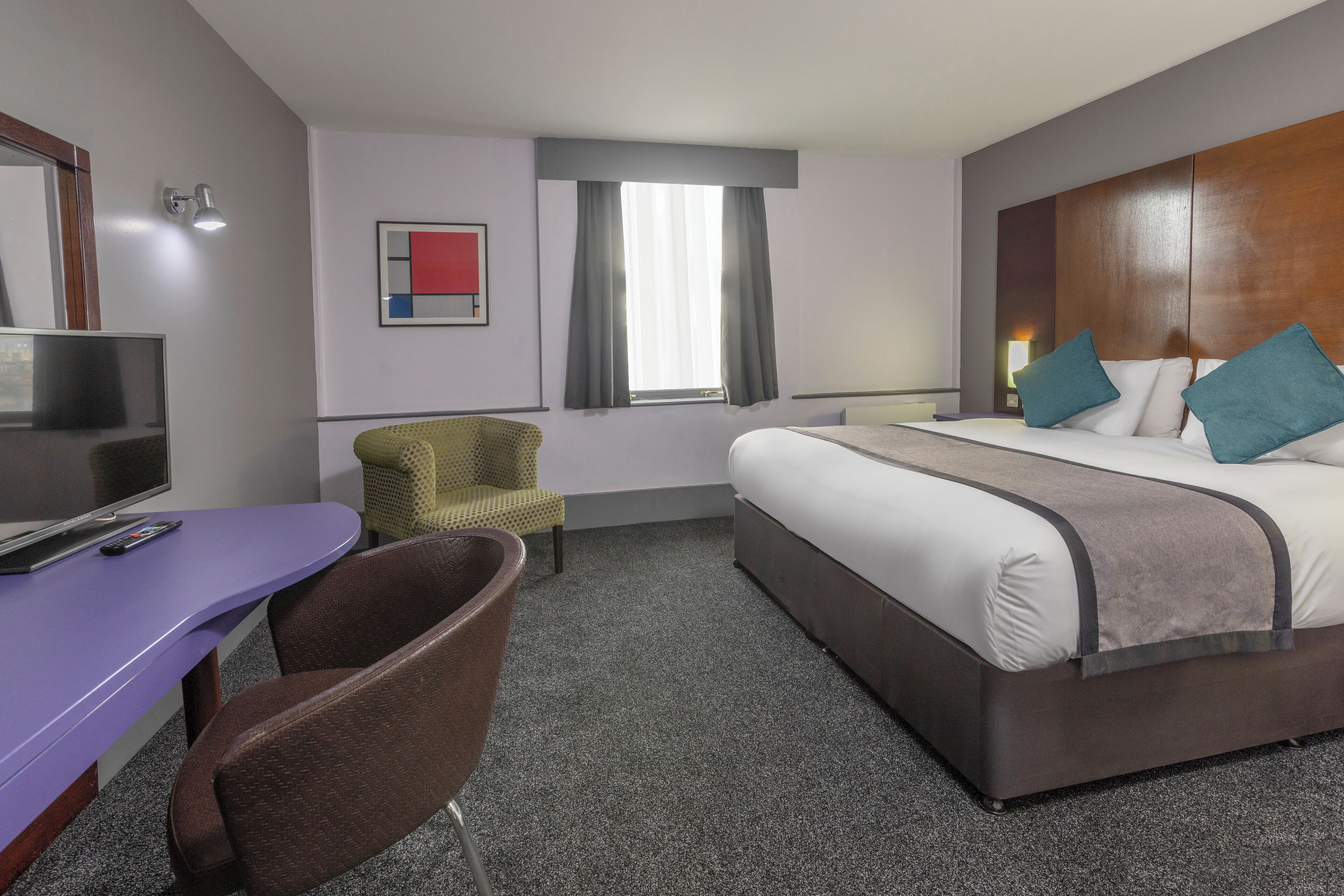 A hotel room with a bed, a desk with a chair, a television, and a window with curtains.
