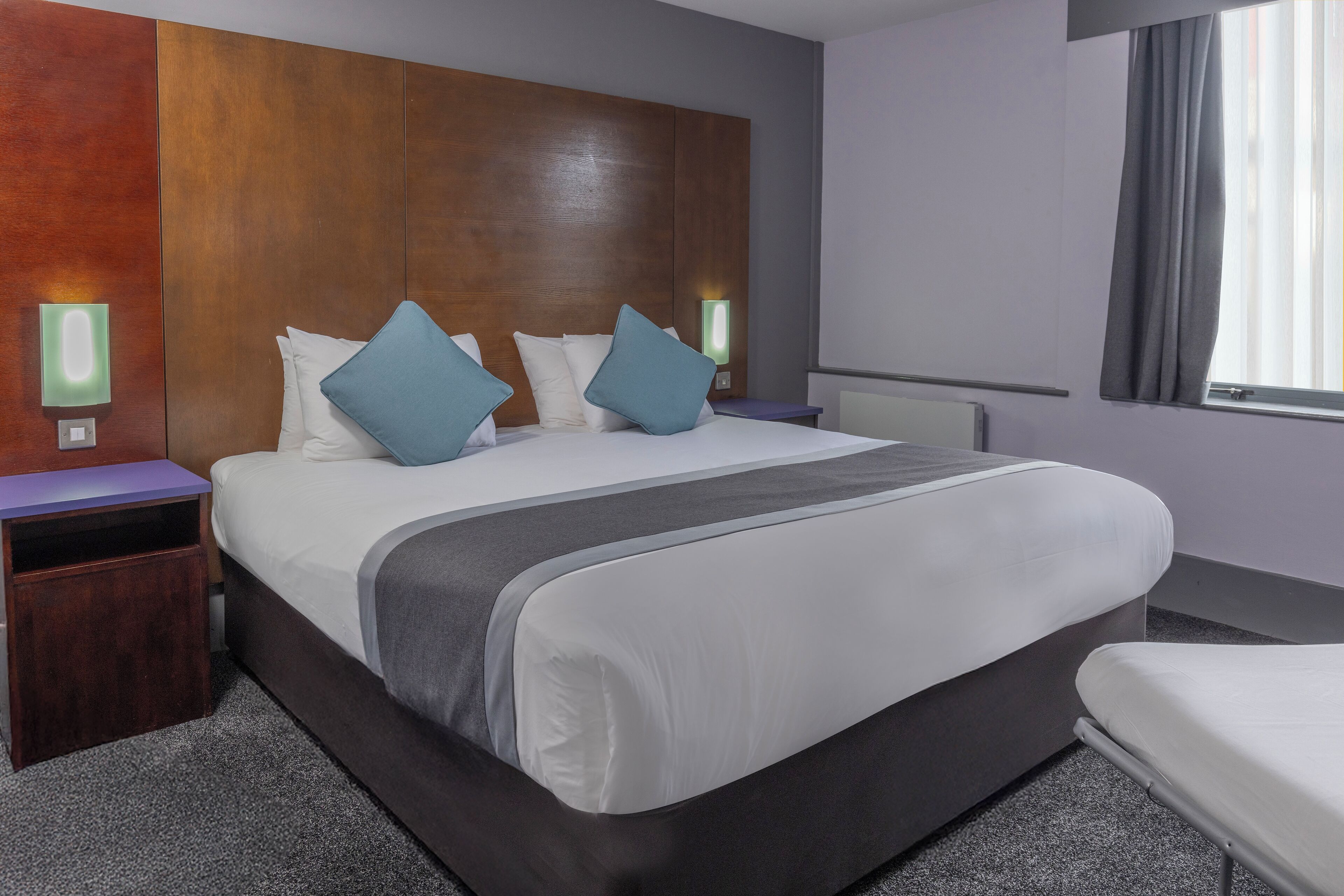 Habitación de hotel con una cama grande, dos mesitas de noche, una lámpara de pared y una ventana con cortinas.