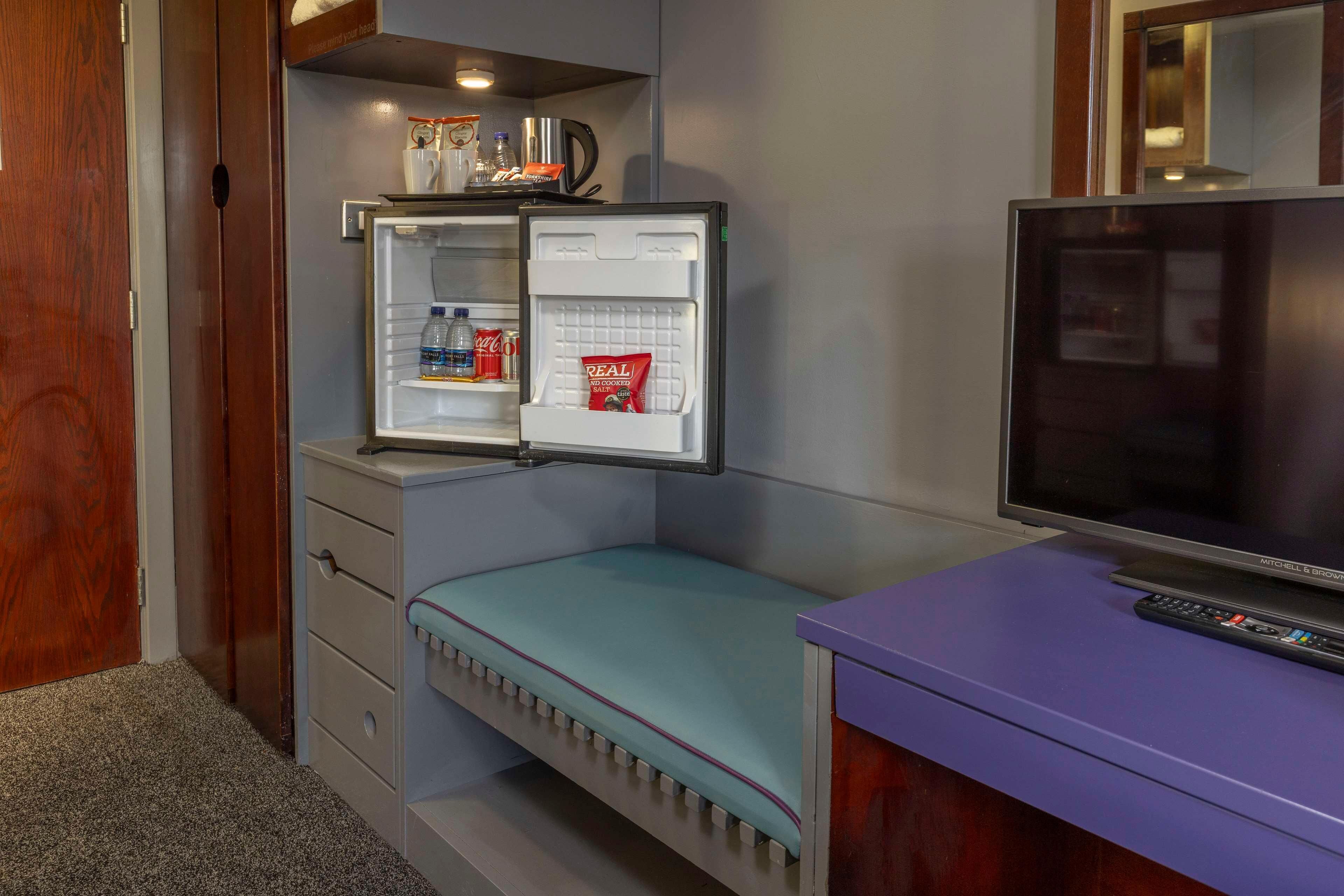Una habitación compacta con refrigerador incorporado, televisión sobre un soporte y un pequeño banco.