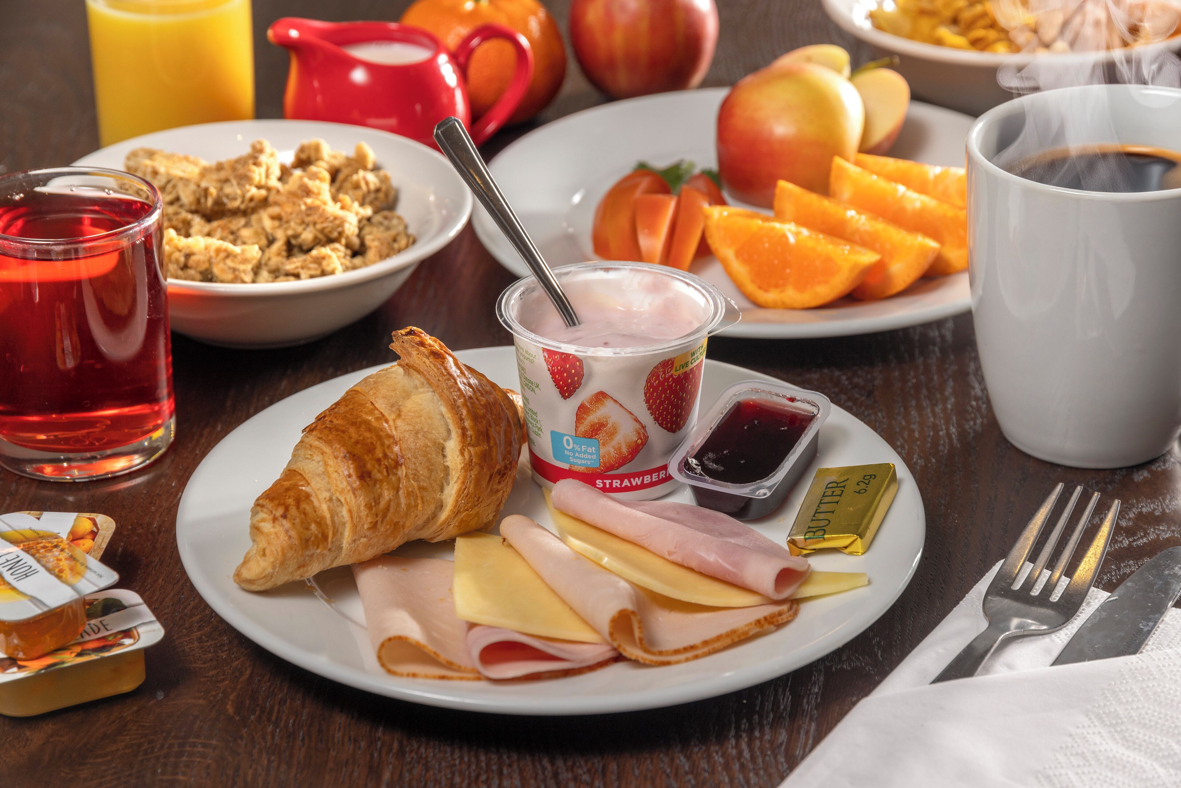 Ontbijtbord met croissant, ham, kaas en boter, vergezeld van een glas rood sap, een kom ontbijtgranen en een schaal fruit.
