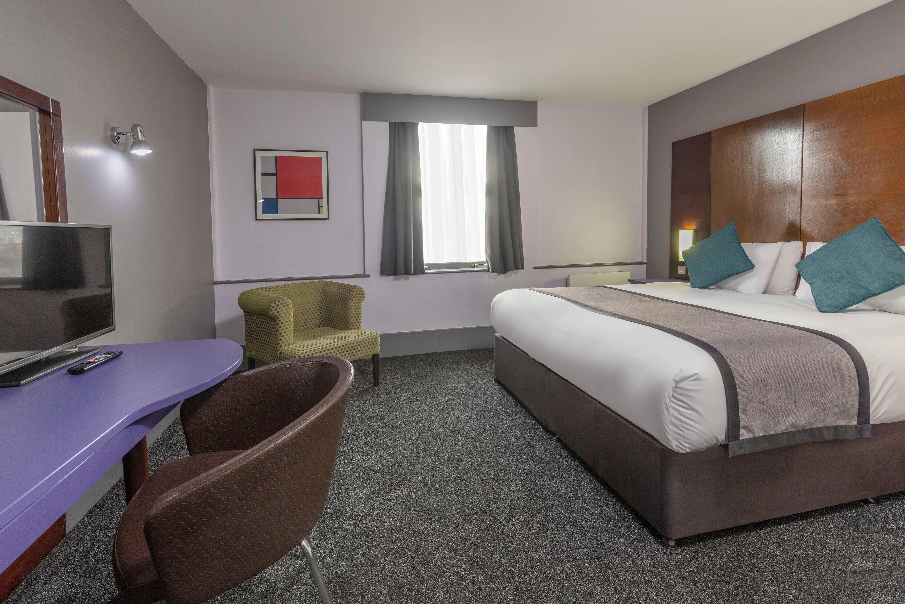 A hotel room with a bed, a desk with a chair, a television, and a window with curtains.