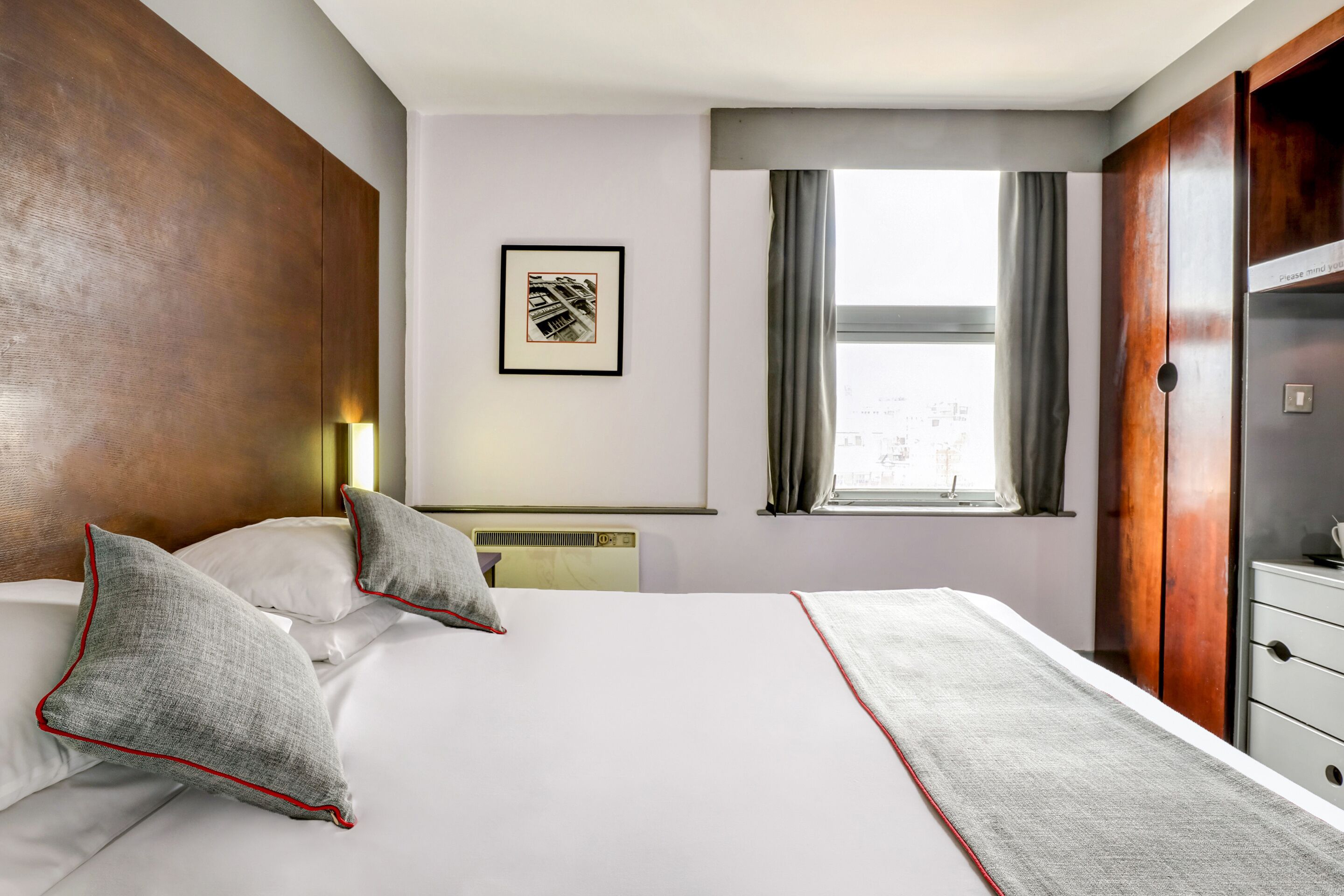 A hotel room with a large bed, a framed picture on the wall, a window with curtains, and a wooden wardrobe.