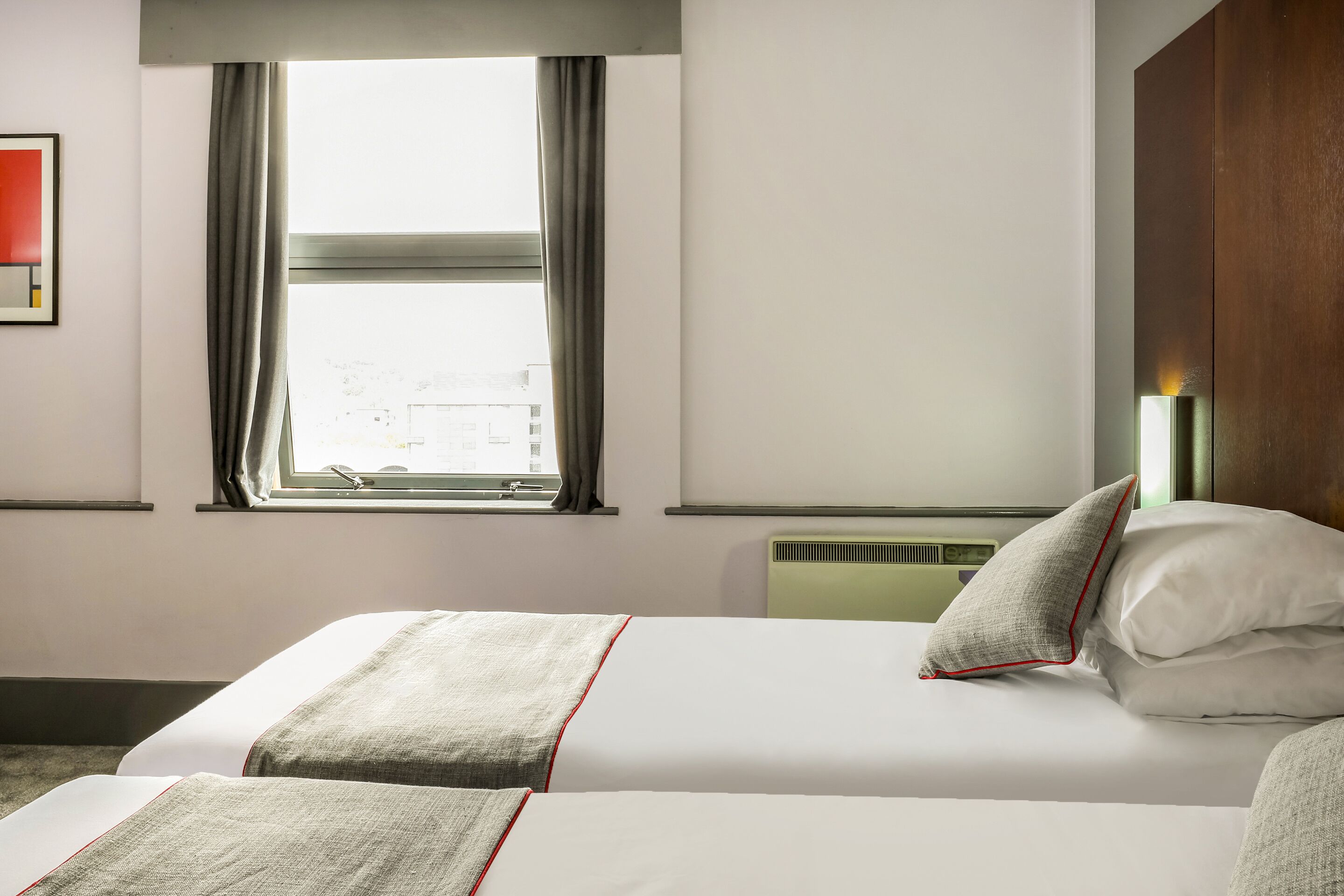 A hotel room with two beds, a window with curtains, a wall-mounted light, and a radiator.