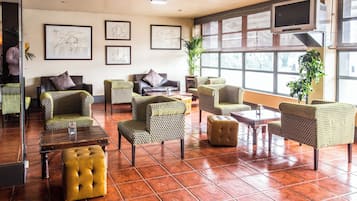 Un vestíbulo amplio con varias zonas de estar, incluyendo sofás y sillones, una mesa de centro y un suelo de baldosas. Se observan obras de arte enmarcadas en la pared y ventanas grandes que proporcionan luz natural.