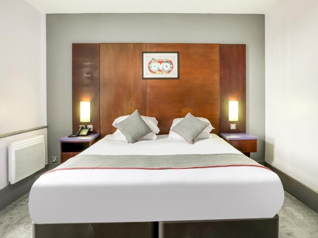 A hotel room with a large bed, two bedside tables, a headboard with a framed picture, and wall-mounted lamps.