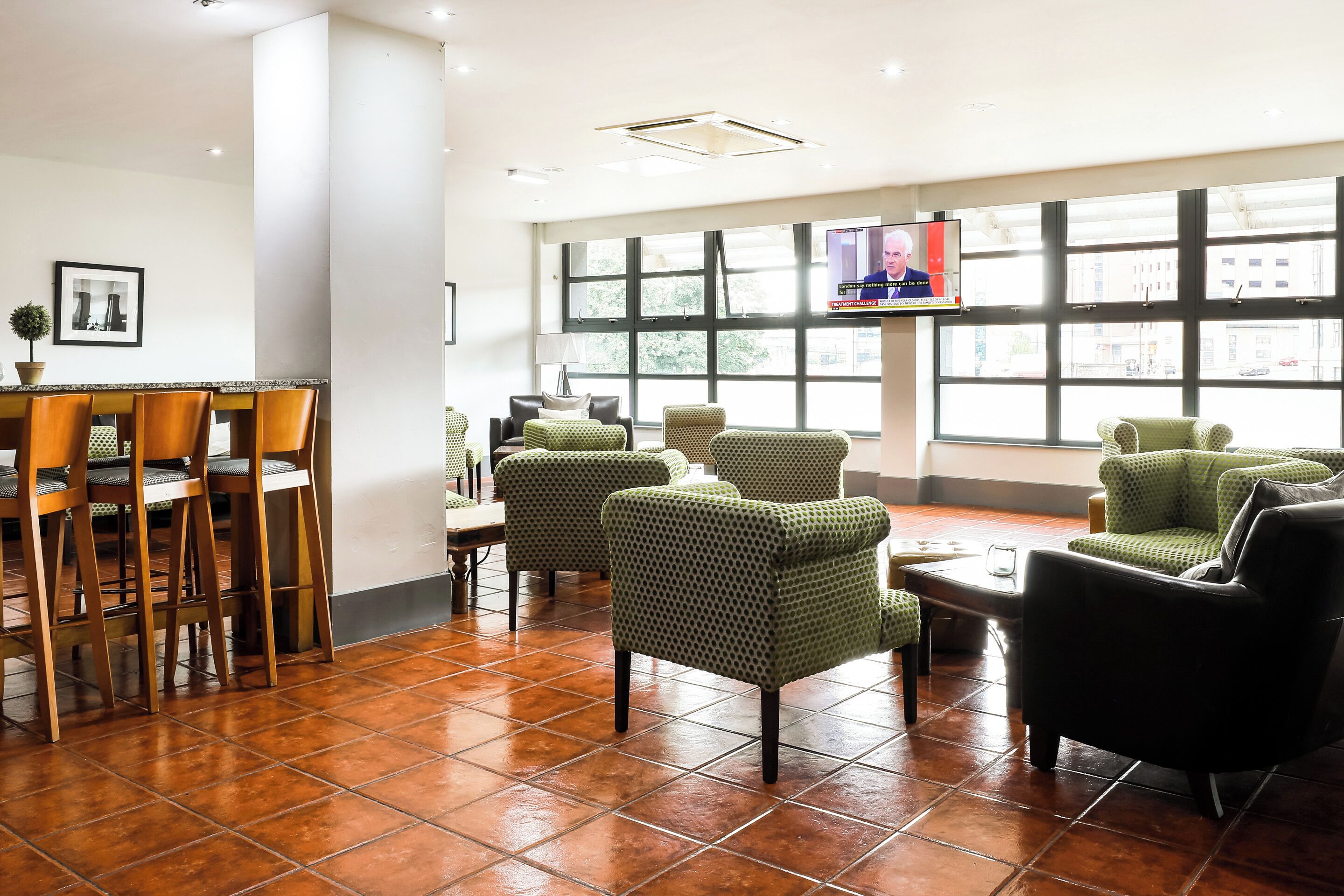 A hotel lobby with a dining area, green and black upholstered chairs, a television screen, and large windows.