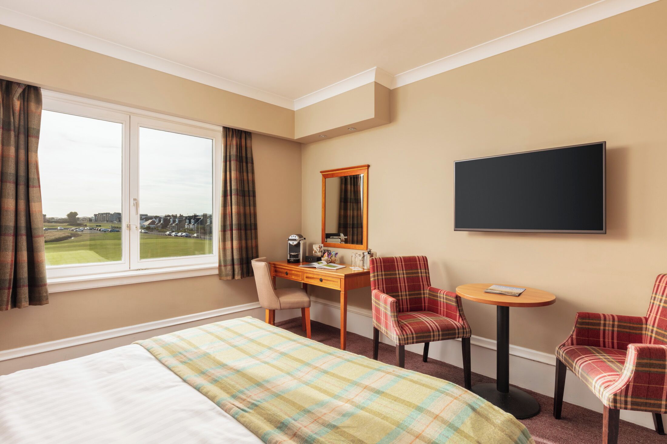 A hotel room with a bed, a desk, two chairs, a small table, a flat-screen TV, and a window with curtains.