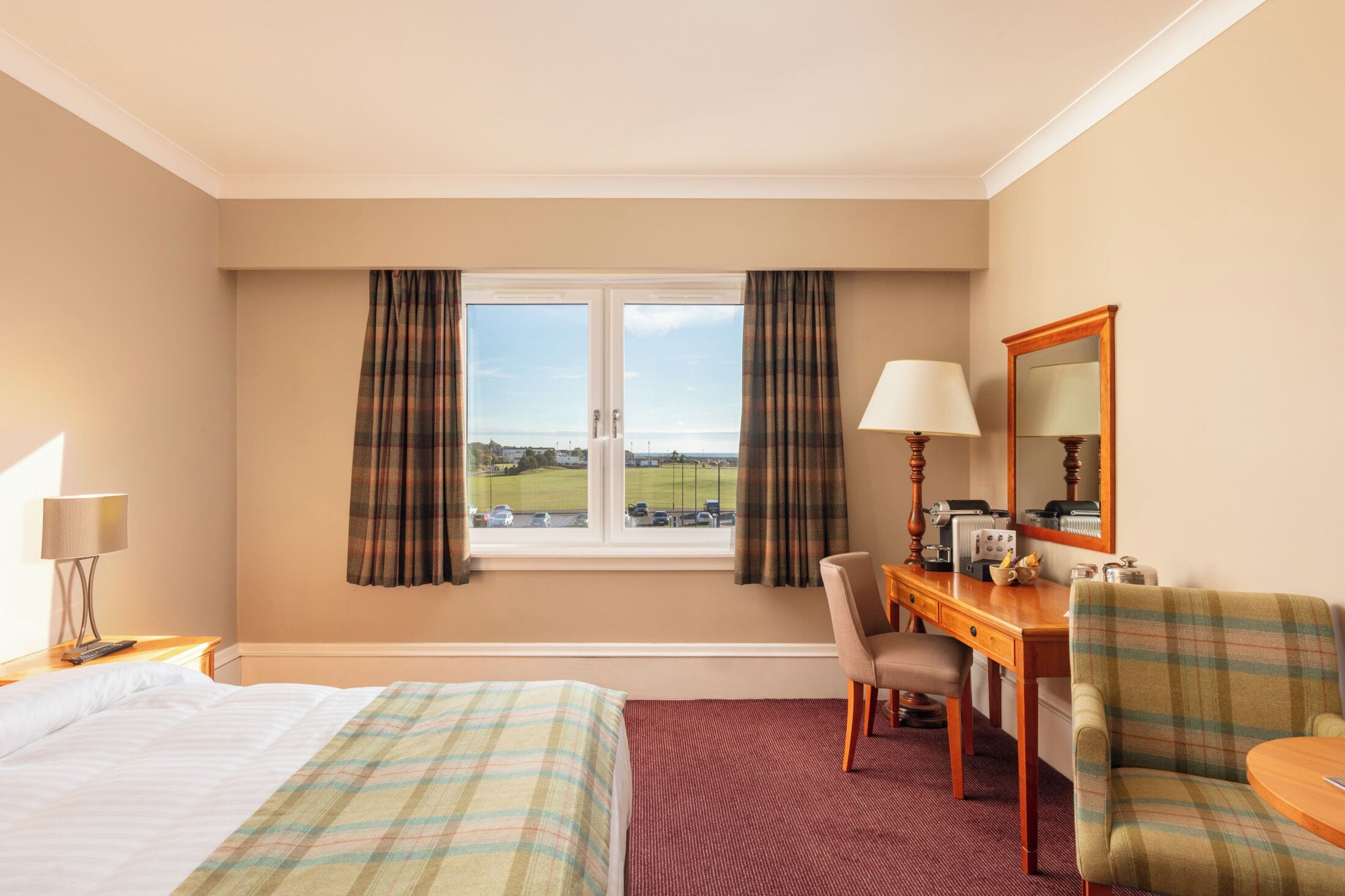 A hotel room with a bed, a desk with a chair, a lamp, a mirror, and a window with curtains.