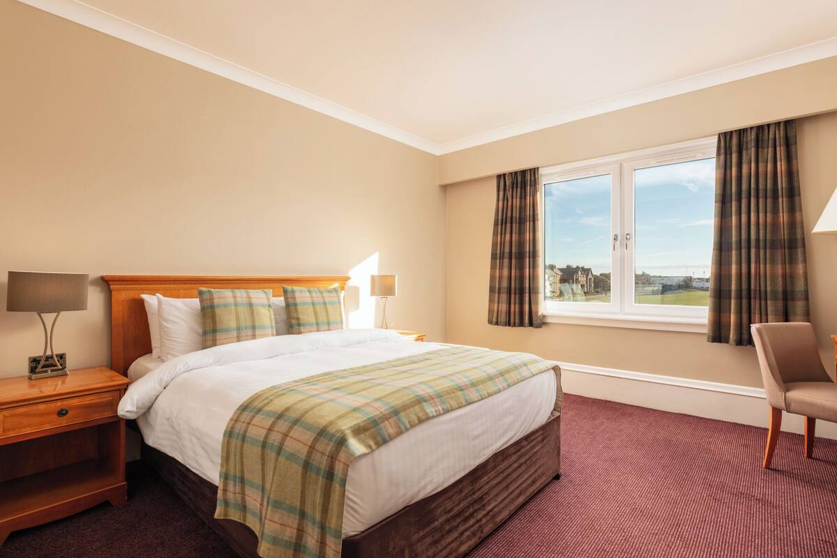 A hotel room with a large bed, a wooden nightstand, a chair, and a window with curtains.