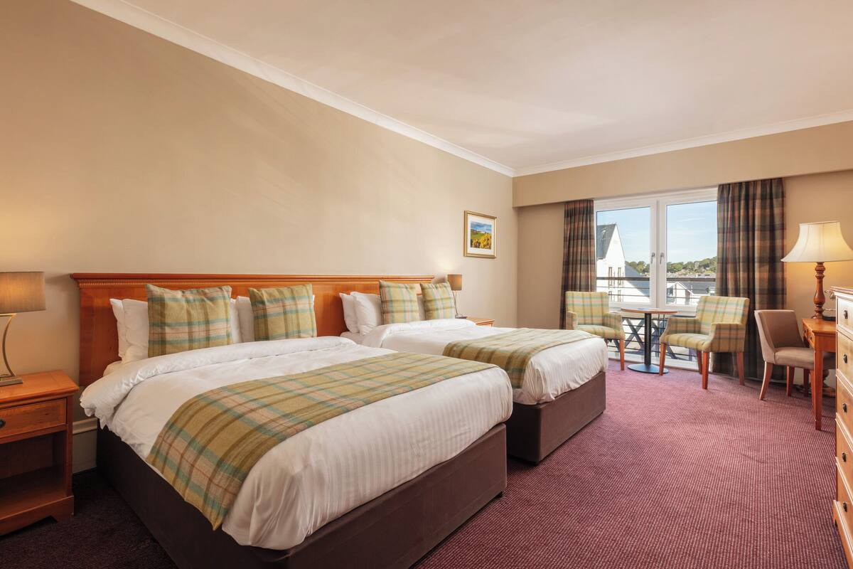 Habitación de hotel con dos camas, una mesita, sillas y vistas al exterior.