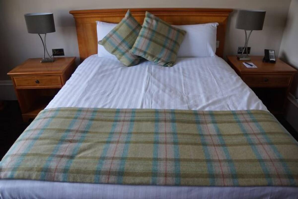 Habitación de hotel con una cama, dos mesitas de noche, una lámpara sobre cada una y una colcha en la cama.
