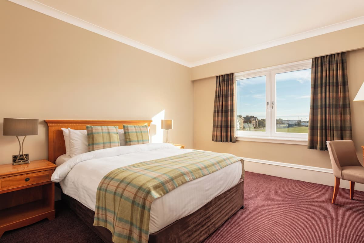 Habitación de hotel con una cama grande, una mesita de noche de madera, una silla y una ventana con cortinas.