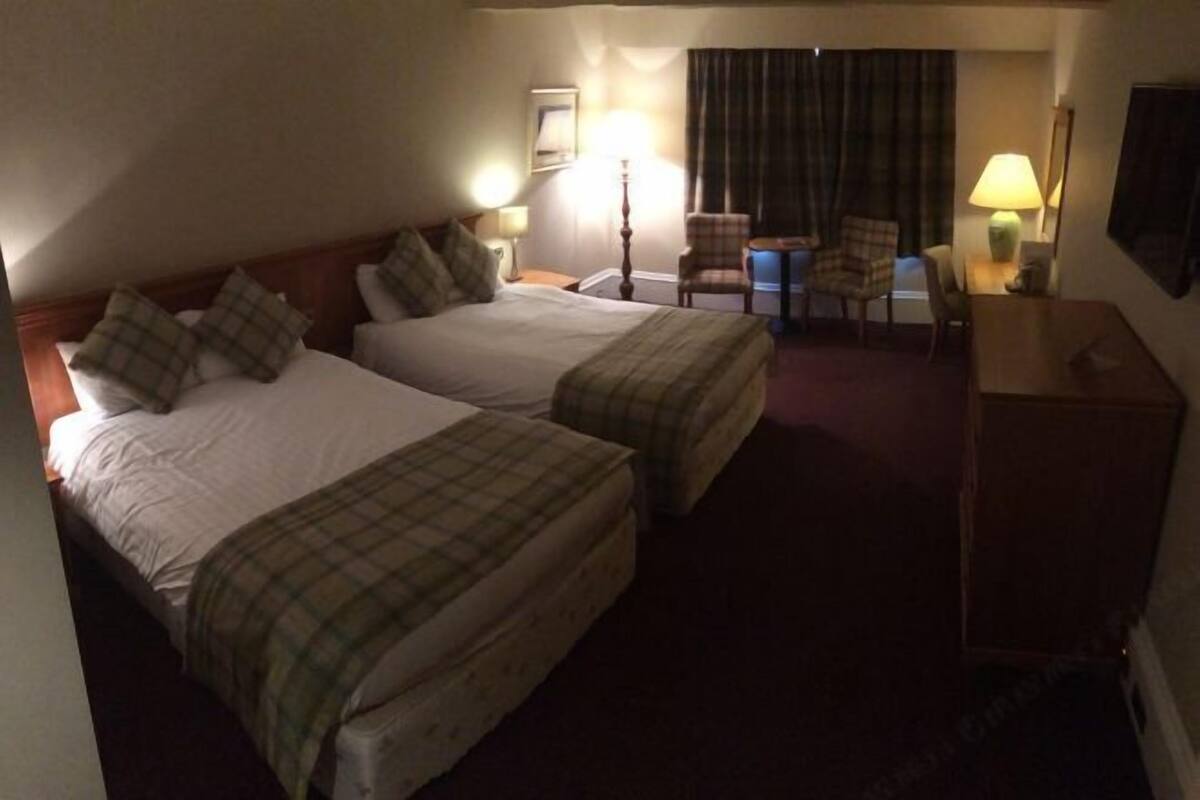 A hotel room with two beds, a desk, a chair, and a lamp.