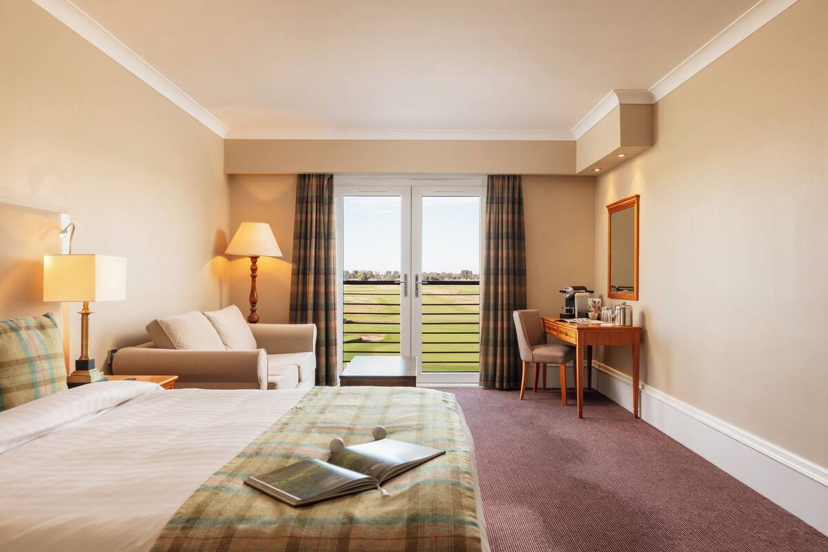 A hotel room with a bed, a desk, a chair, a lamp, and a view through the window.