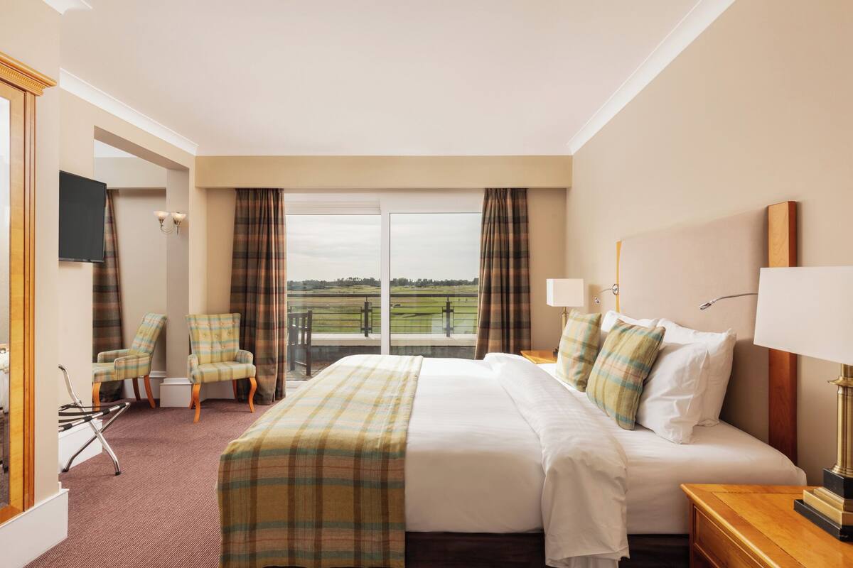 Habitación de hotel con una cama grande, dos sillones, un televisor y un balcón con vistas al campo.