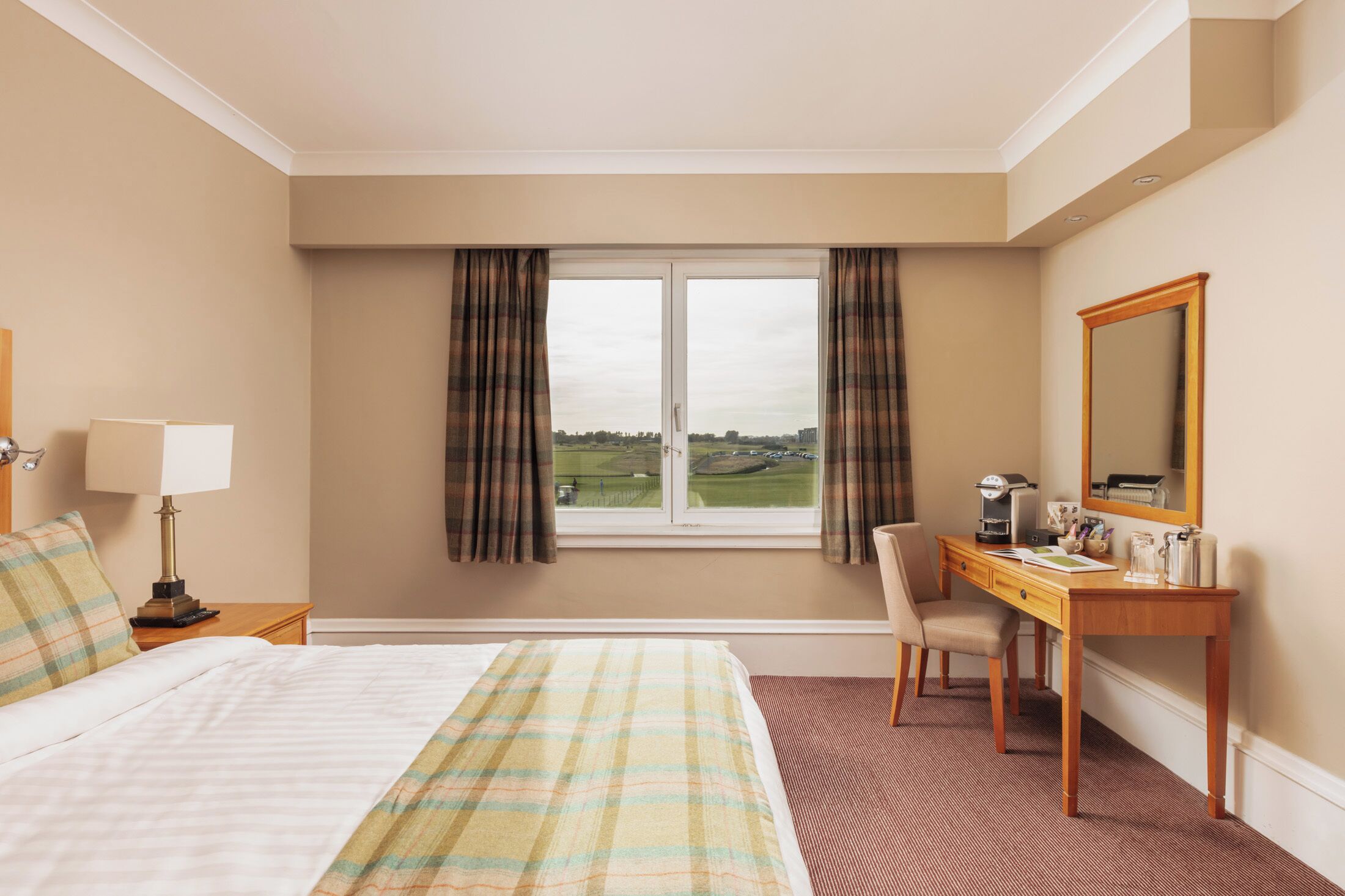 A hotel room with a bed, a desk, a chair, a lamp, a mirror, and a window with curtains.