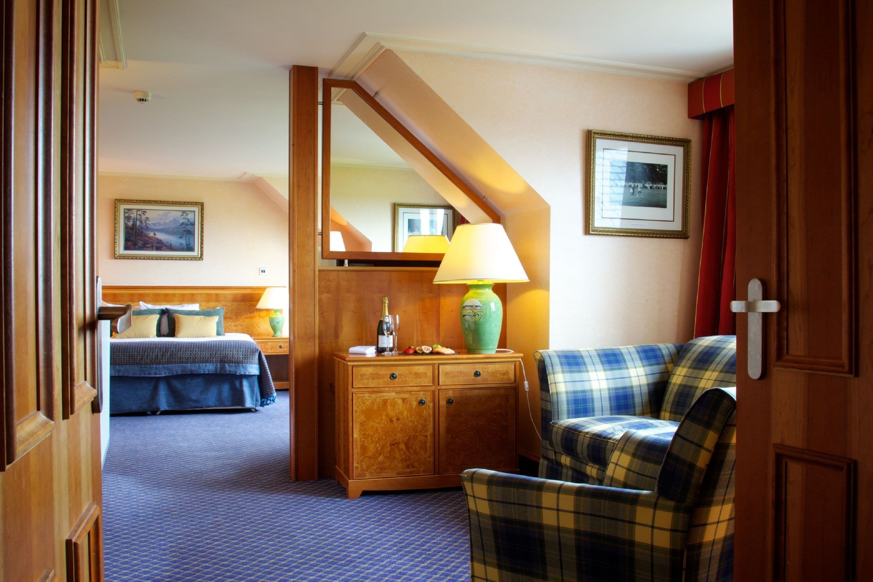 Une chambre d’hôtel avec un lit, une armoire en bois, un fauteuil à motifs écossais et un miroir qui reflète la pièce.