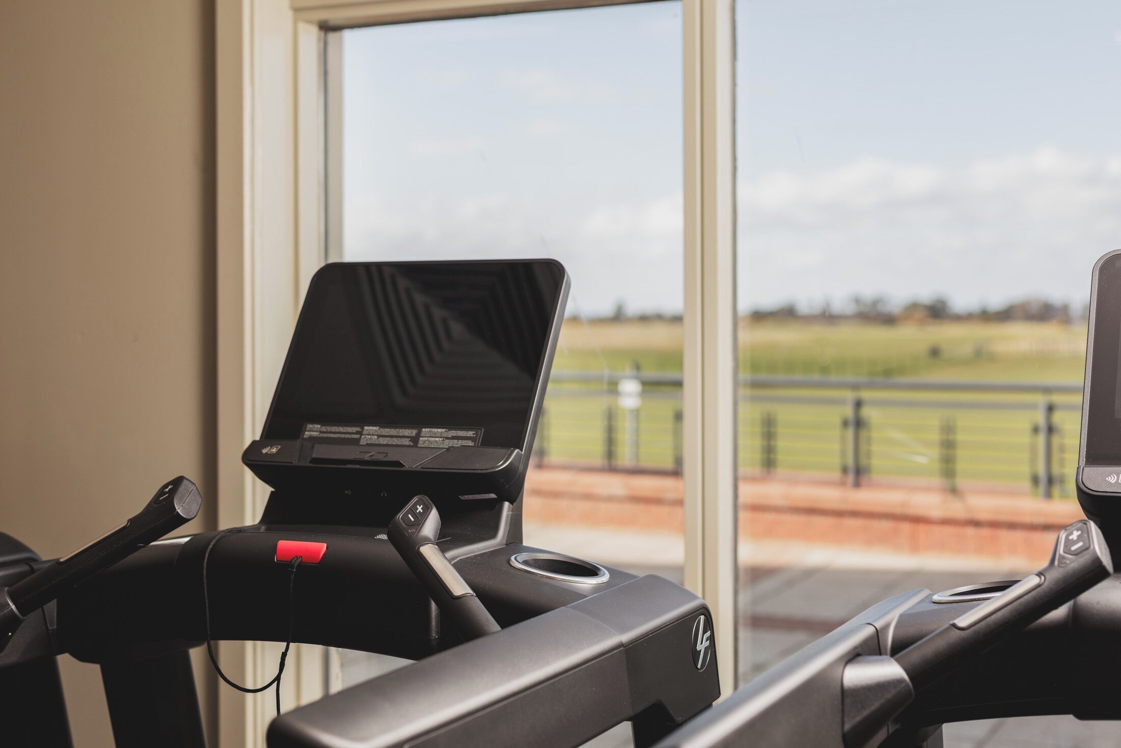 A treadmill with a digital display, positioned near a window with a view of a green field and a fence.