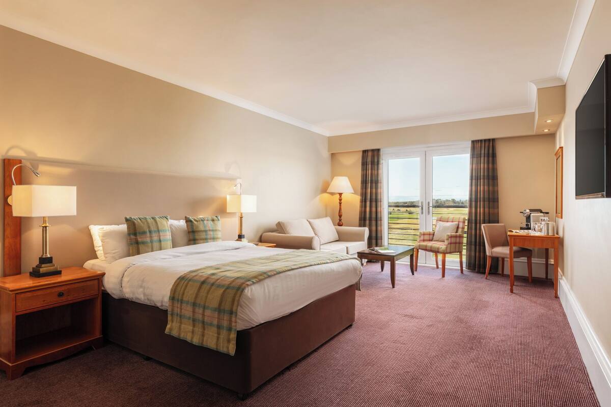 Habitación de hotel con una cama grande, dos mesitas de noche con lámparas, un sofá, una pequeña mesa y una silla. Cuenta con una ventana con cortinas y vistas al exterior.