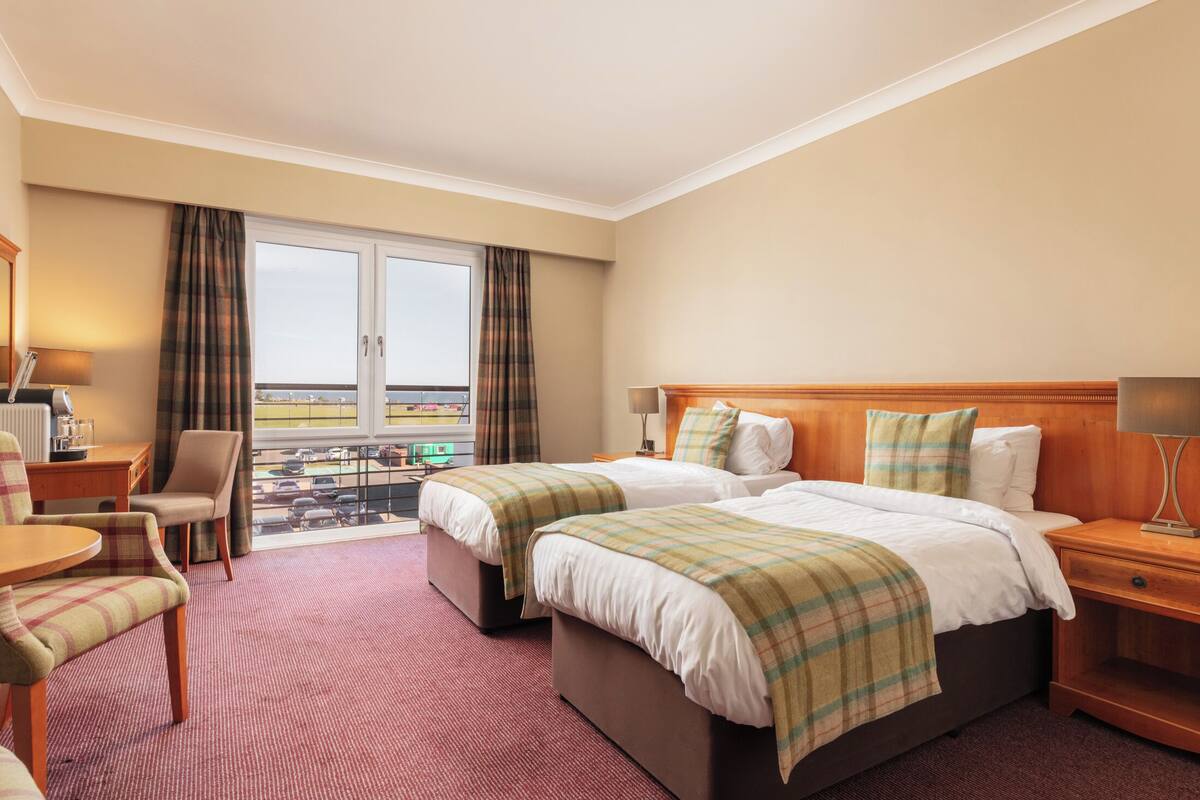 A hotel room with two beds, a desk, a chair, and a balcony with a view.