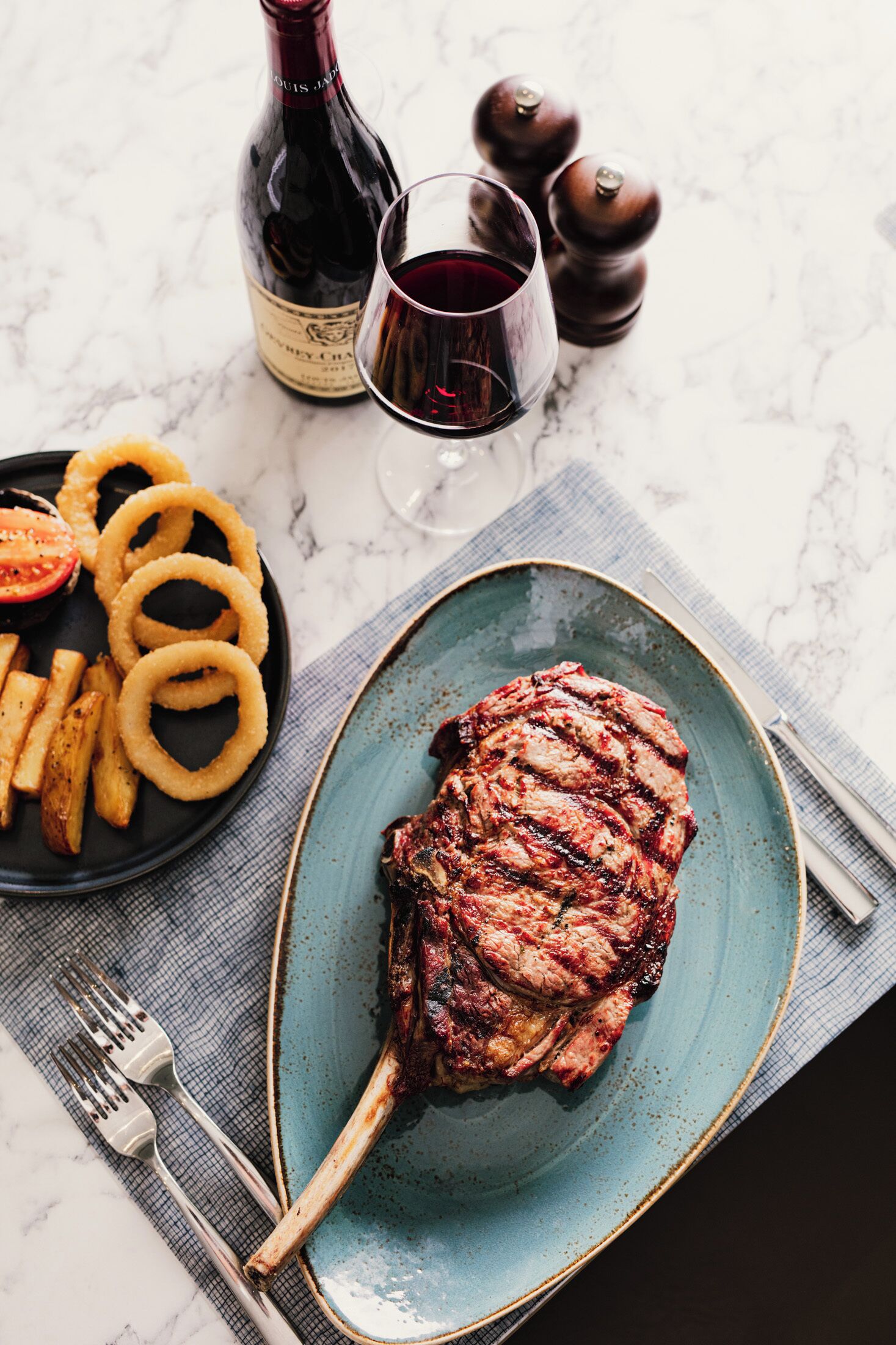 A plate with grilled steak, onion rings, and fries on a blue plate, with a bottle of wine and a glass of red wine in the background.