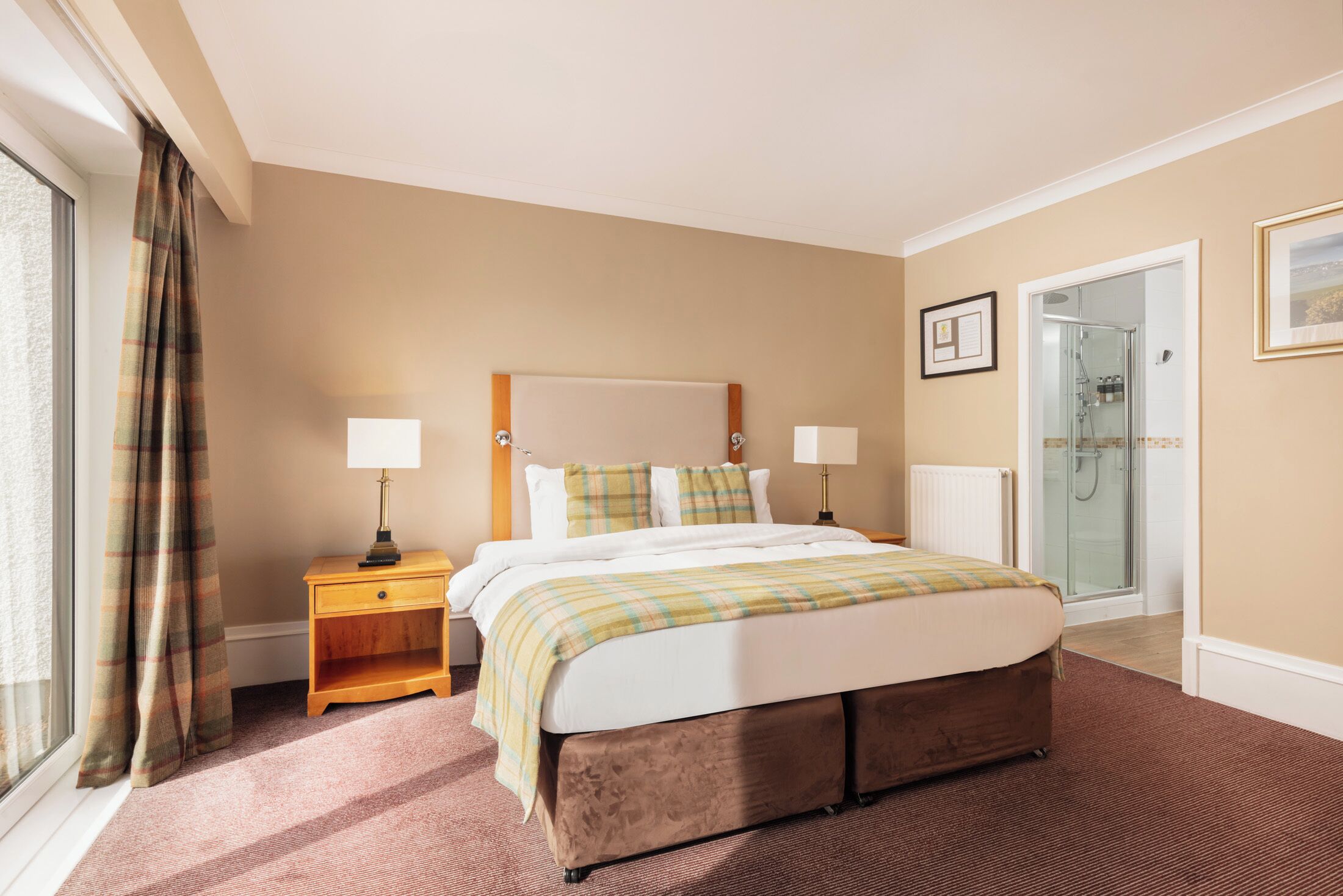 A hotel room with a large bed, bedside lamps, a nightstand, a glass shower door, and a framed picture on the wall.