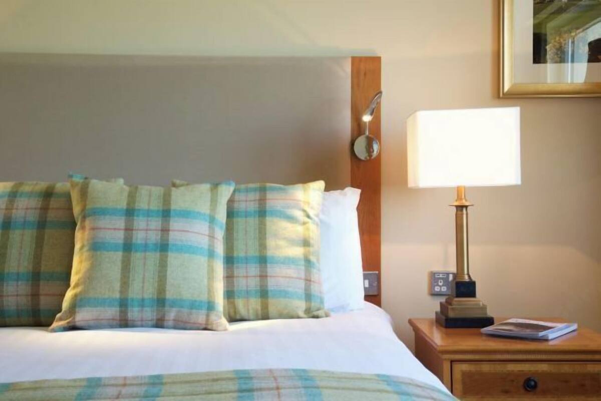 Una cama bien hecha con ropa de cama escocesa, una lámpara de noche y una mesita con un libro.
