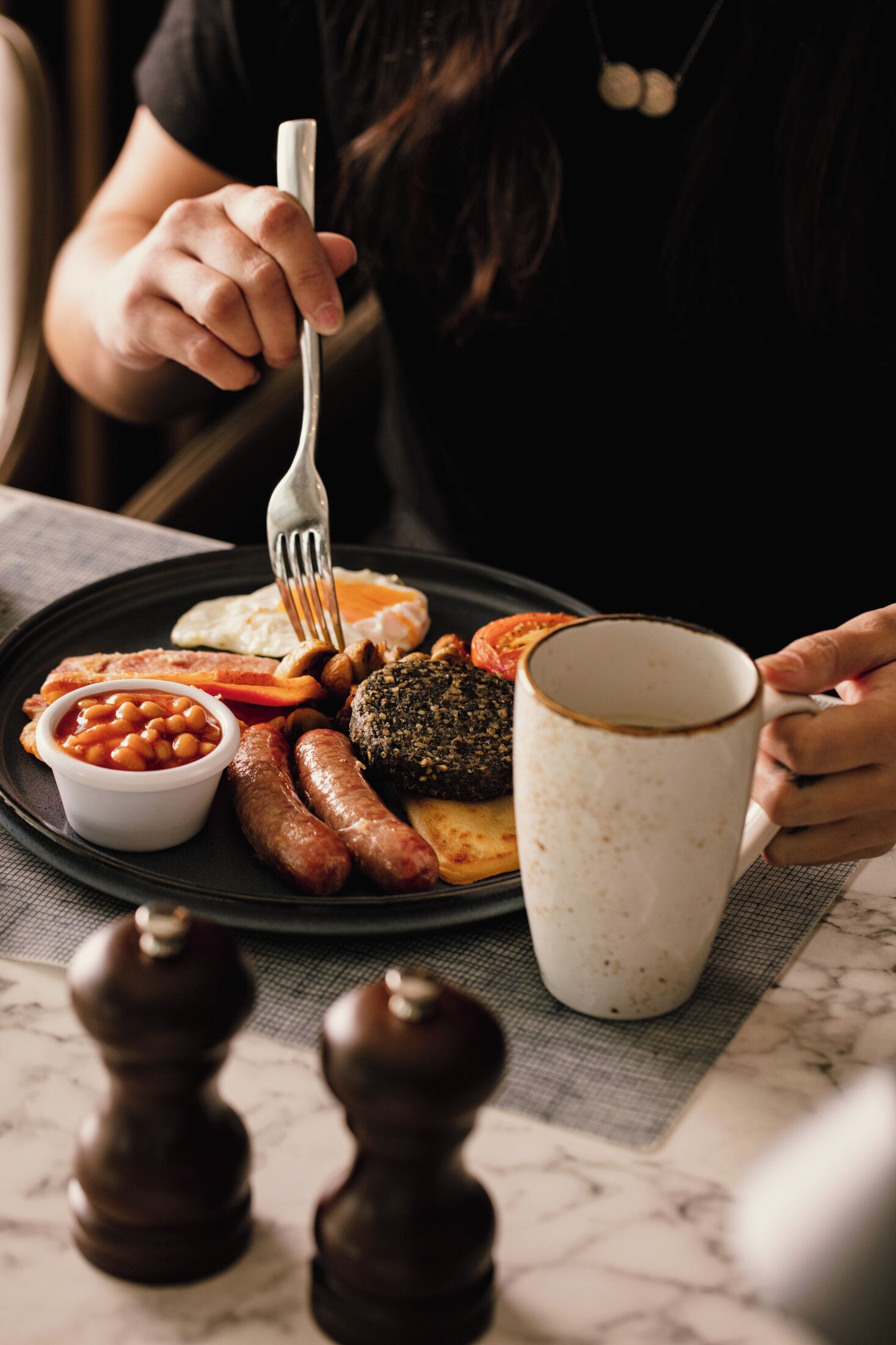 A plate of breakfast food including sausages, eggs, and beans, with a mug and pepper shakers on a table.