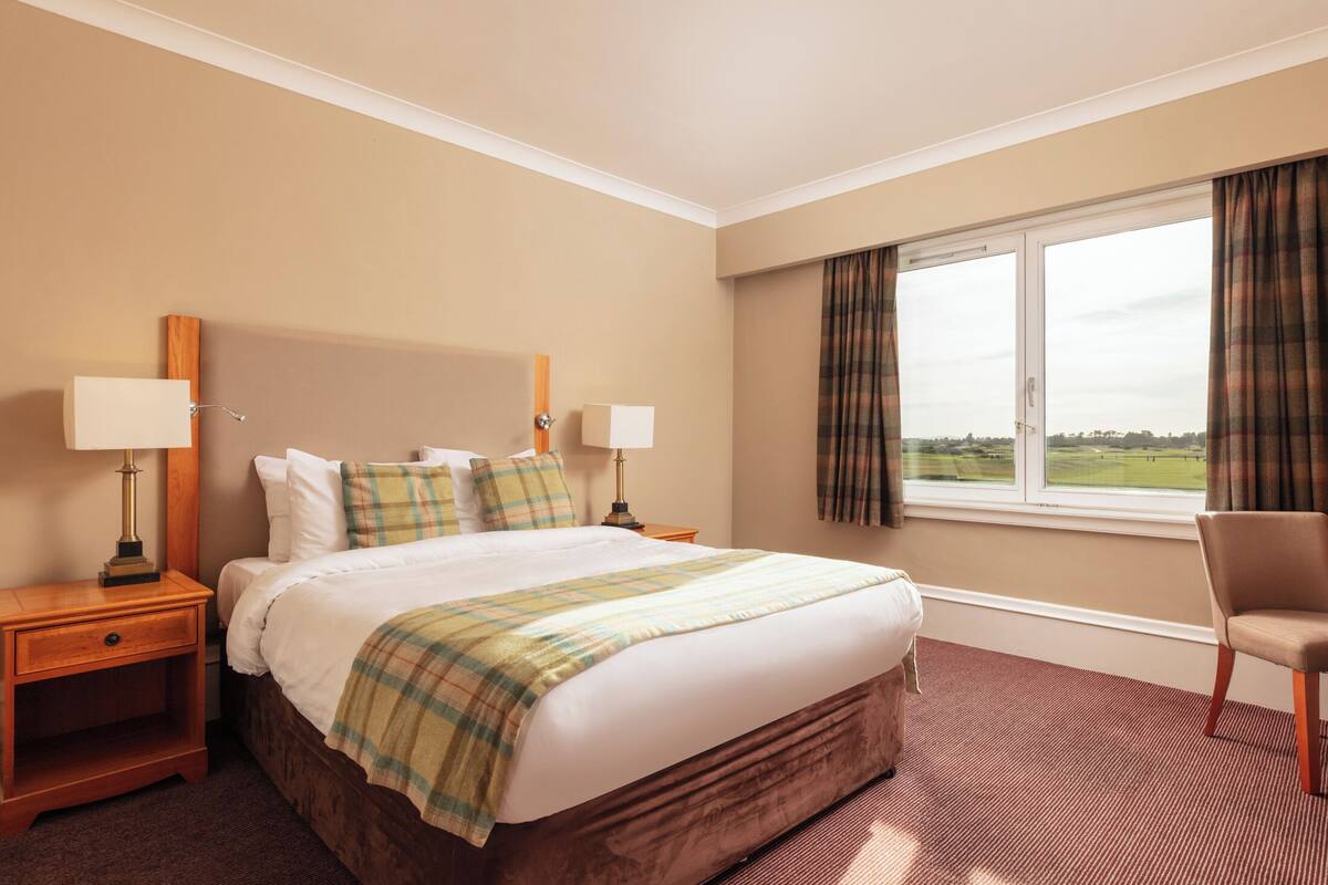 Habitación de hotel con una cama grande, dos mesitas de noche con lámparas, una silla, una ventana con cortinas y vistas al exterior.