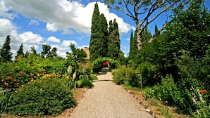 Un camí al jardí que condueix a un edifici amb teulada vermella, envoltat de plantes i arbres diversos.
