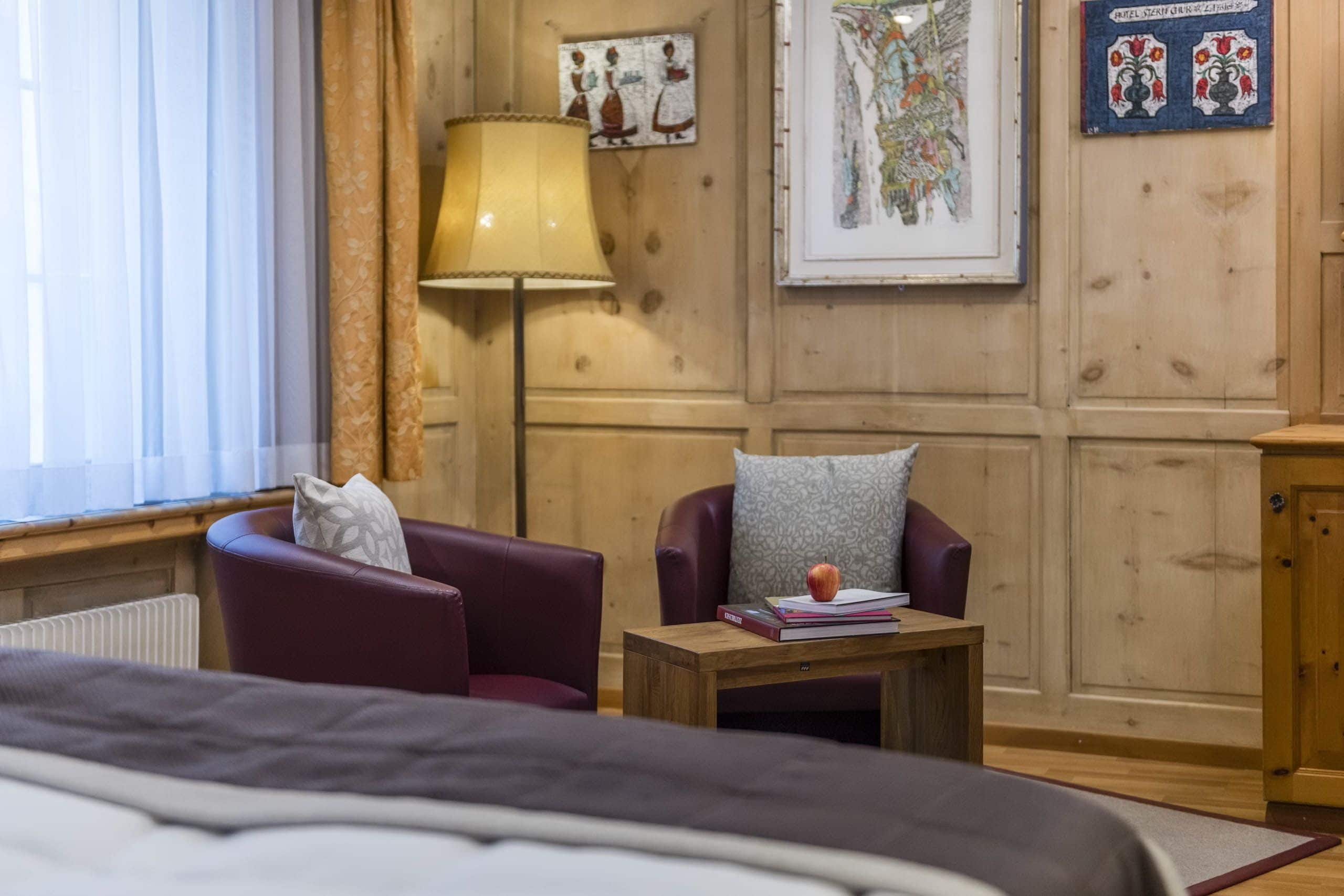 Ein gemütliches Zimmer mit einem Bett, einem Nachttisch aus Holz, einer Lampe, einem Sessel und einem kleinen Tisch mit Büchern und einem Apfel.