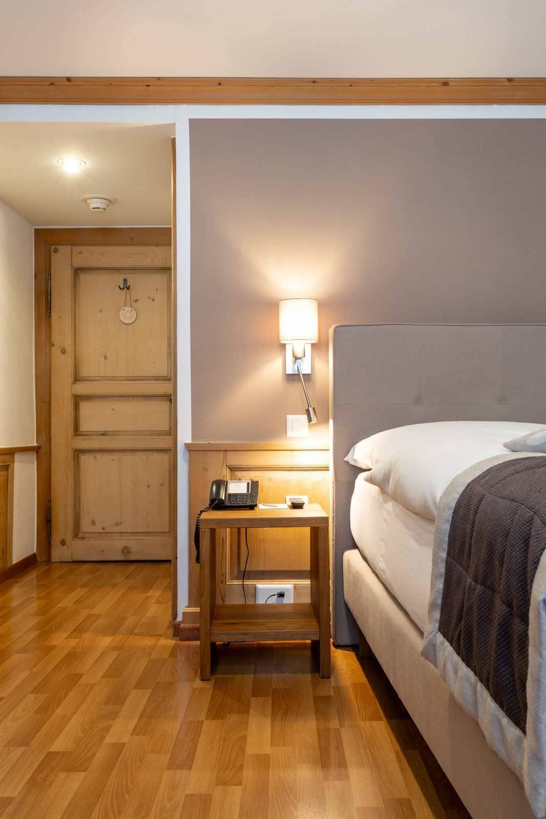 A bedroom with a wooden floor, a bed with white and dark bedding, a bedside table with a lamp, and a wooden door.