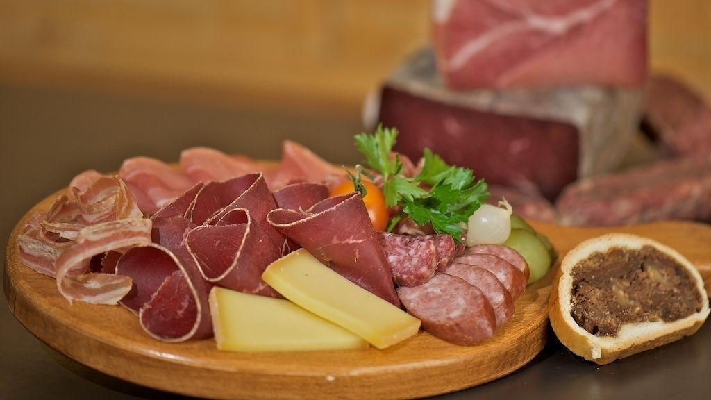 A wooden board with various types of cured meats, including salami and prosciutto, along with a slice of bread and some vegetables.