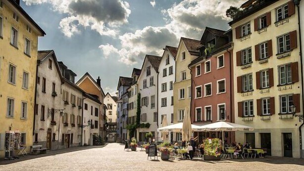 A cobblestone street lined with colorful buildings, a central open space with tables and chairs, and a few people. - Hotel Stern Chur (Chur)