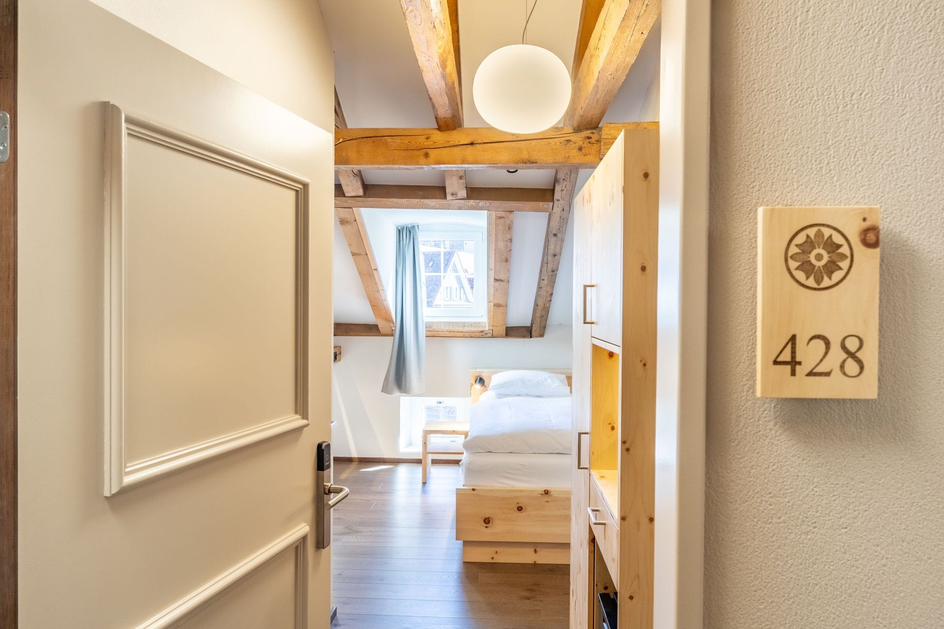 A narrow hallway leading to a bedroom with a wooden ceiling, a bed, and a nightstand. A wooden door with a handle is visible on the left, and a wooden number plaque with the number 428 is on the right wall.