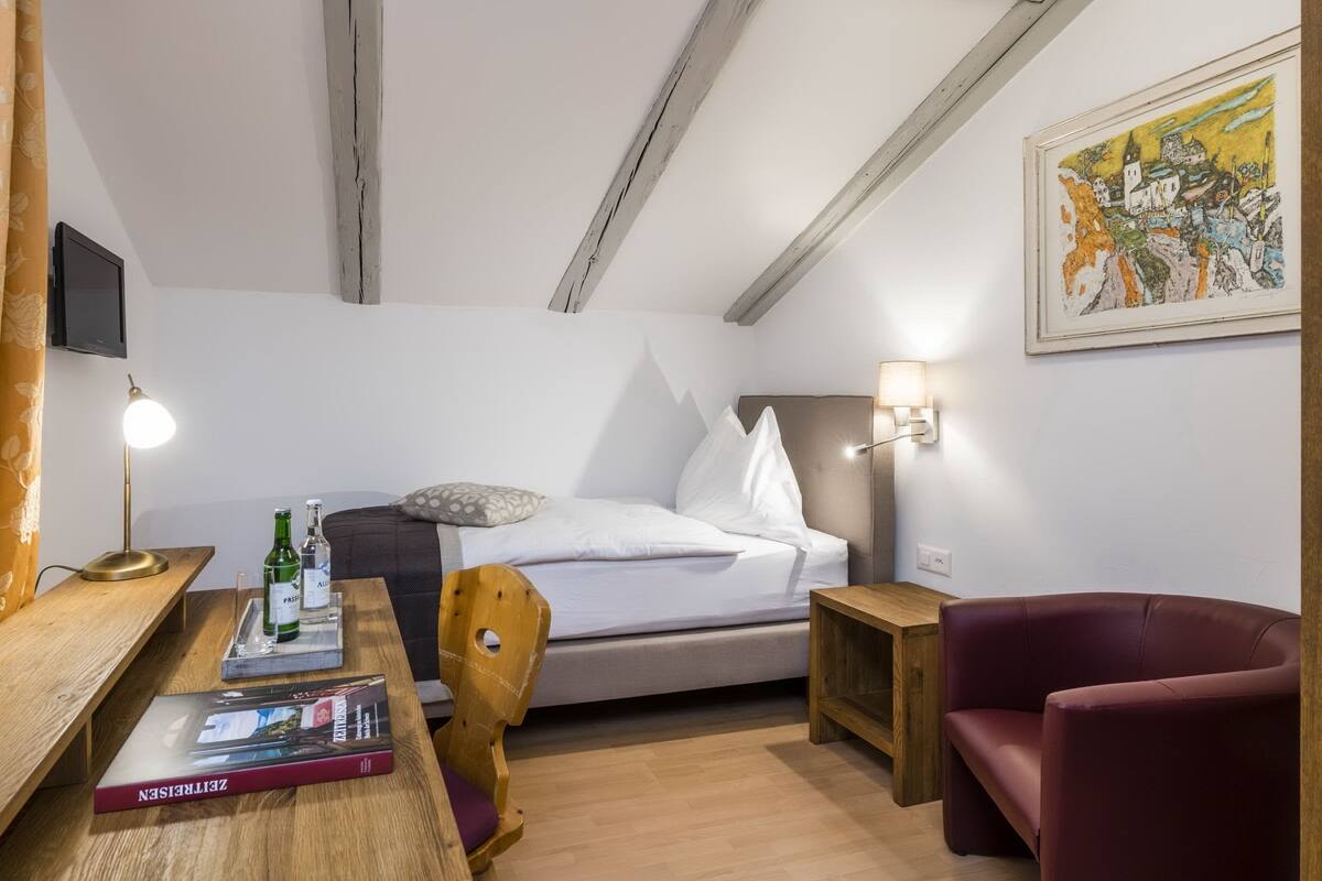 Habitación de hotel con una cama, un escritorio con lámpara, una silla, una mesita y un cuadro en la pared.