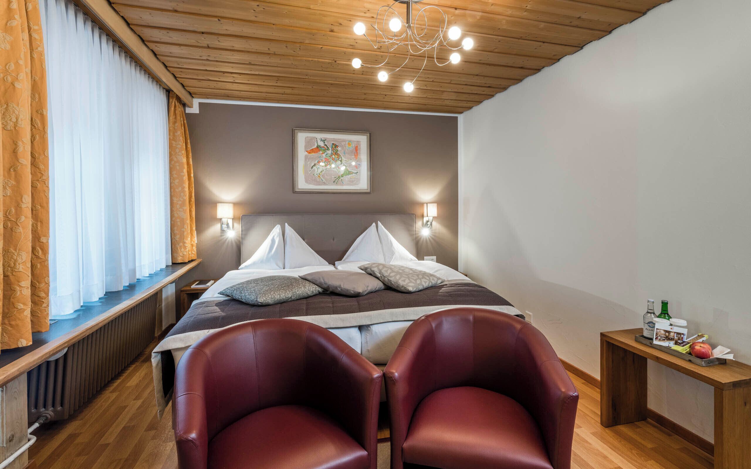 Une chambre d’hôtel avec un plafond en bois, un lit recouvert de draps blancs, deux fauteuils en cuir rouge, une petite table de chevet avec une bouteille et un verre, et un tableau encadré accroché au mur.