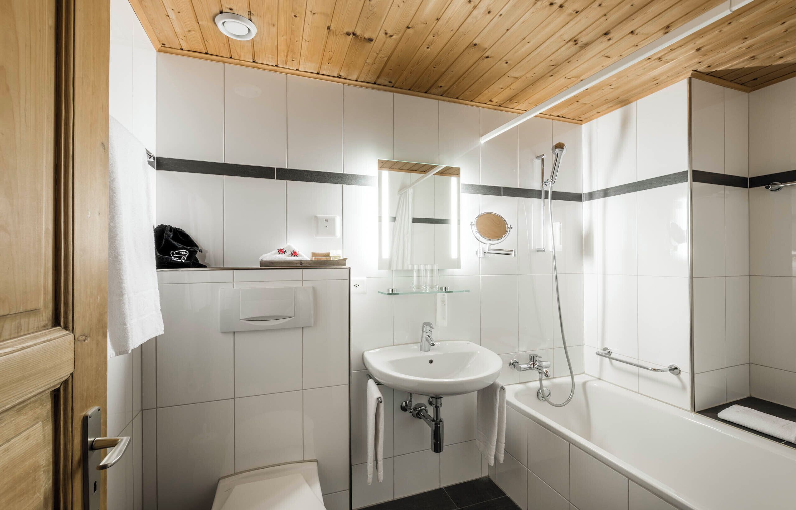 A modern bathroom with a wooden ceiling, white tiles, a sink, a mirror, a shower, and a bathtub.