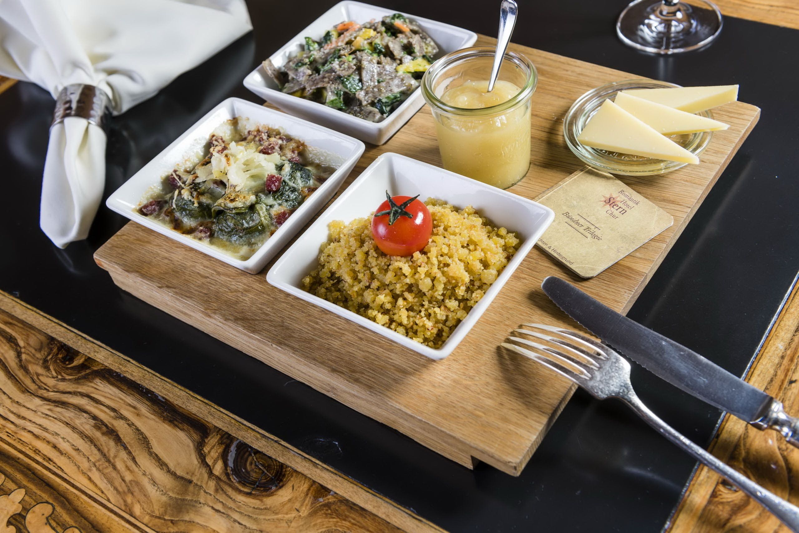 A wooden serving board with three dishes, a glass of juice, and a cheese plate on a black table.