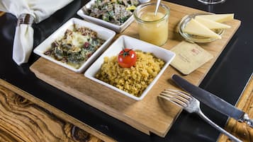 A wooden serving board with three dishes, a glass of juice, and a cheese plate on a black table.
