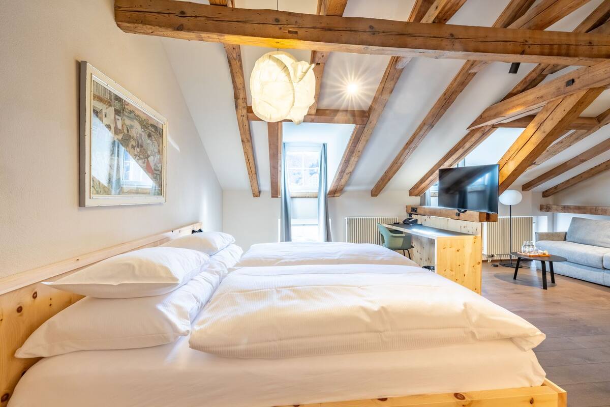 Un dormitorio con techo de madera, una cama con ropa blanca, un escritorio, un televisor y una ventana con cortinas.