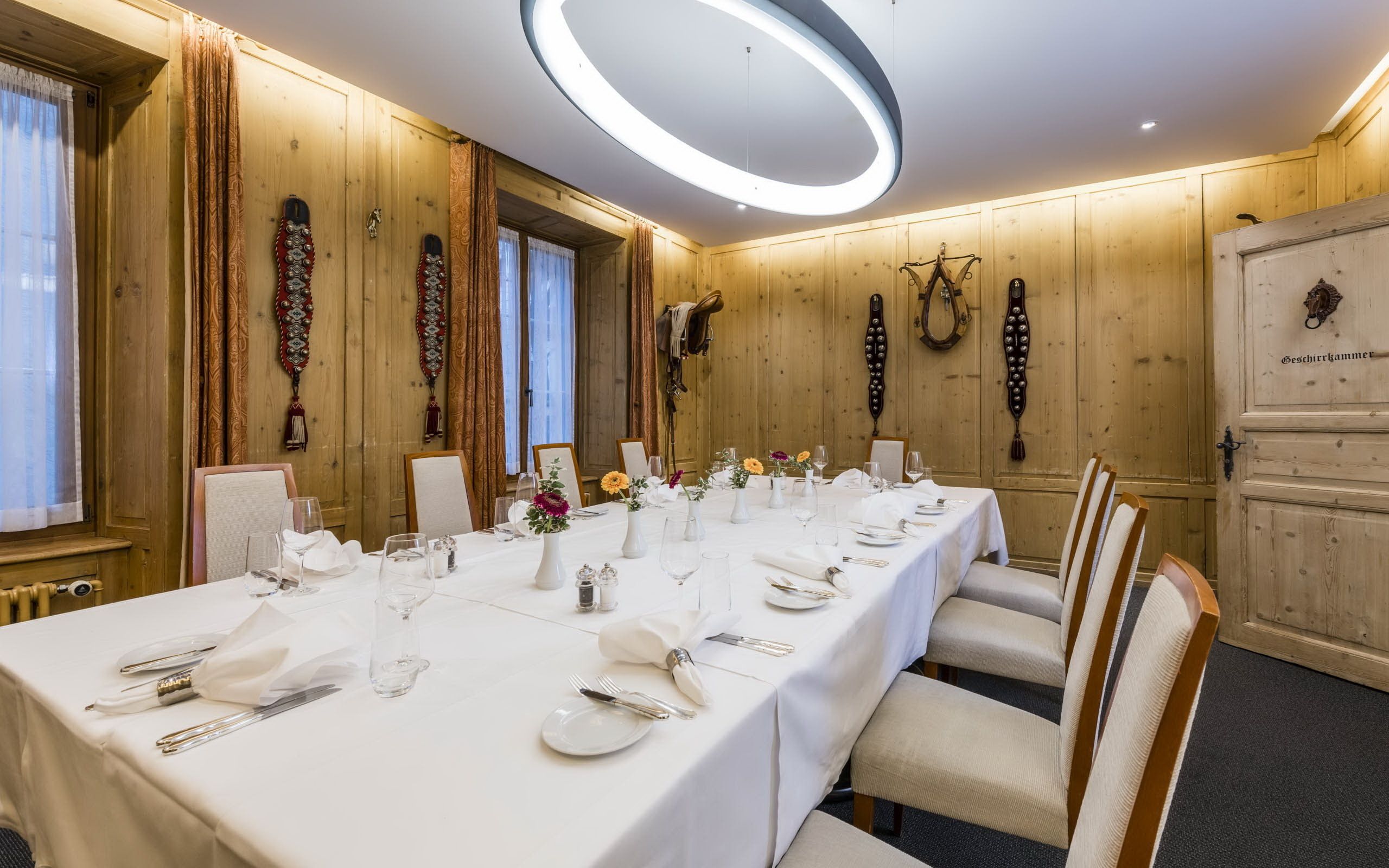 A long dining table set with white tablecloths, glassware, and cutlery, in a room with wooden walls and a modern ceiling light.
