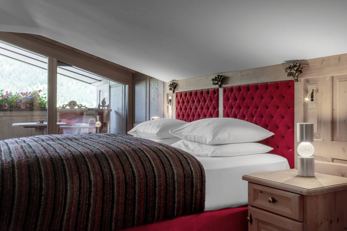 Een slaapkamer met een gestuikt rood hoofdeinde, een nachtkastje met een lamp, en een raam met uitzicht op bloemen.