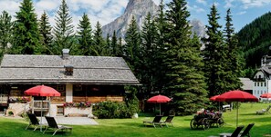 Un chalet avec une façade en bois, des sièges extérieurs ombragés par des parasols rouges et une vue sur les montagnes en arrière-plan.