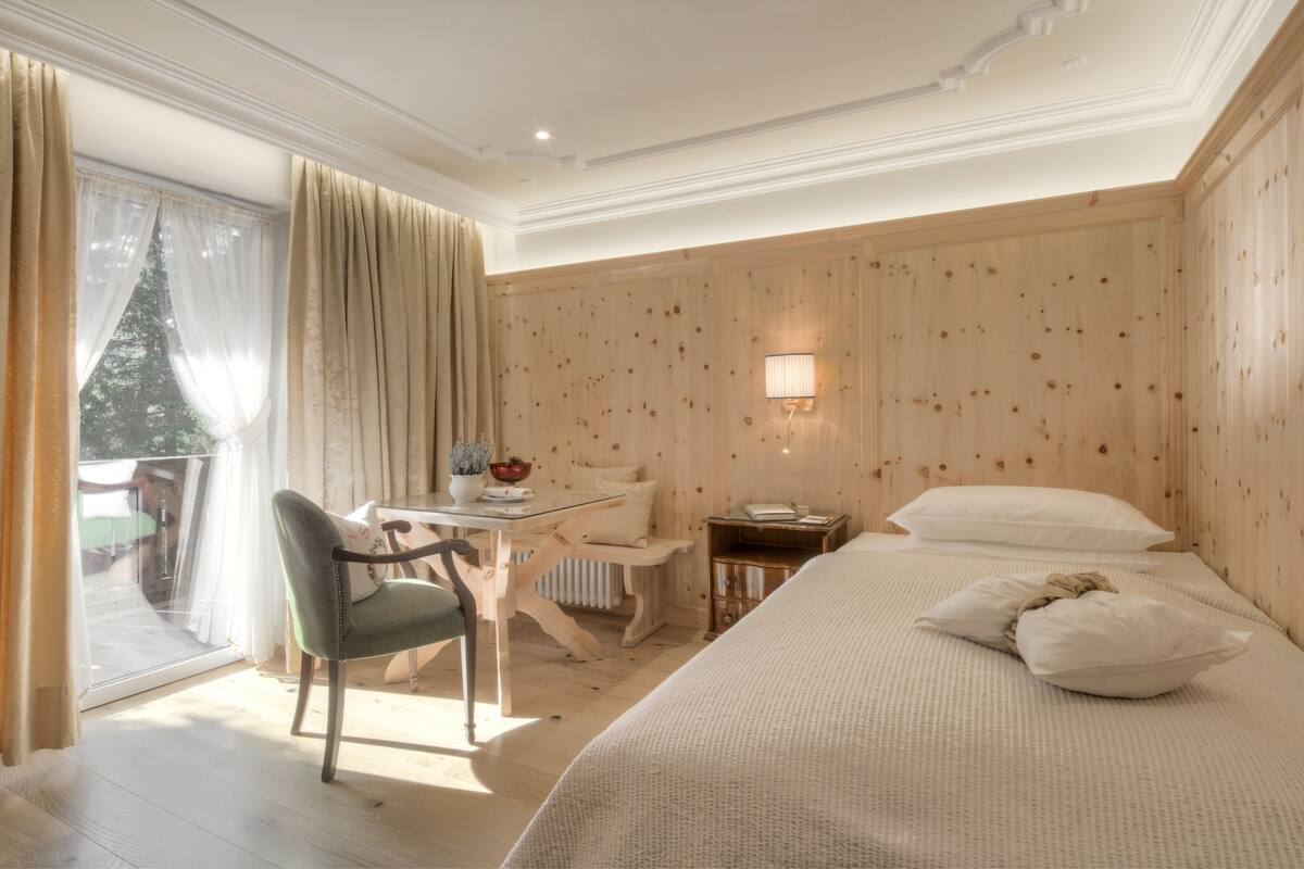 Een hotelkamer met een bed, een bureau, een stoel, een tafeltje, een lamp en uitzicht op een boom door het raam.
