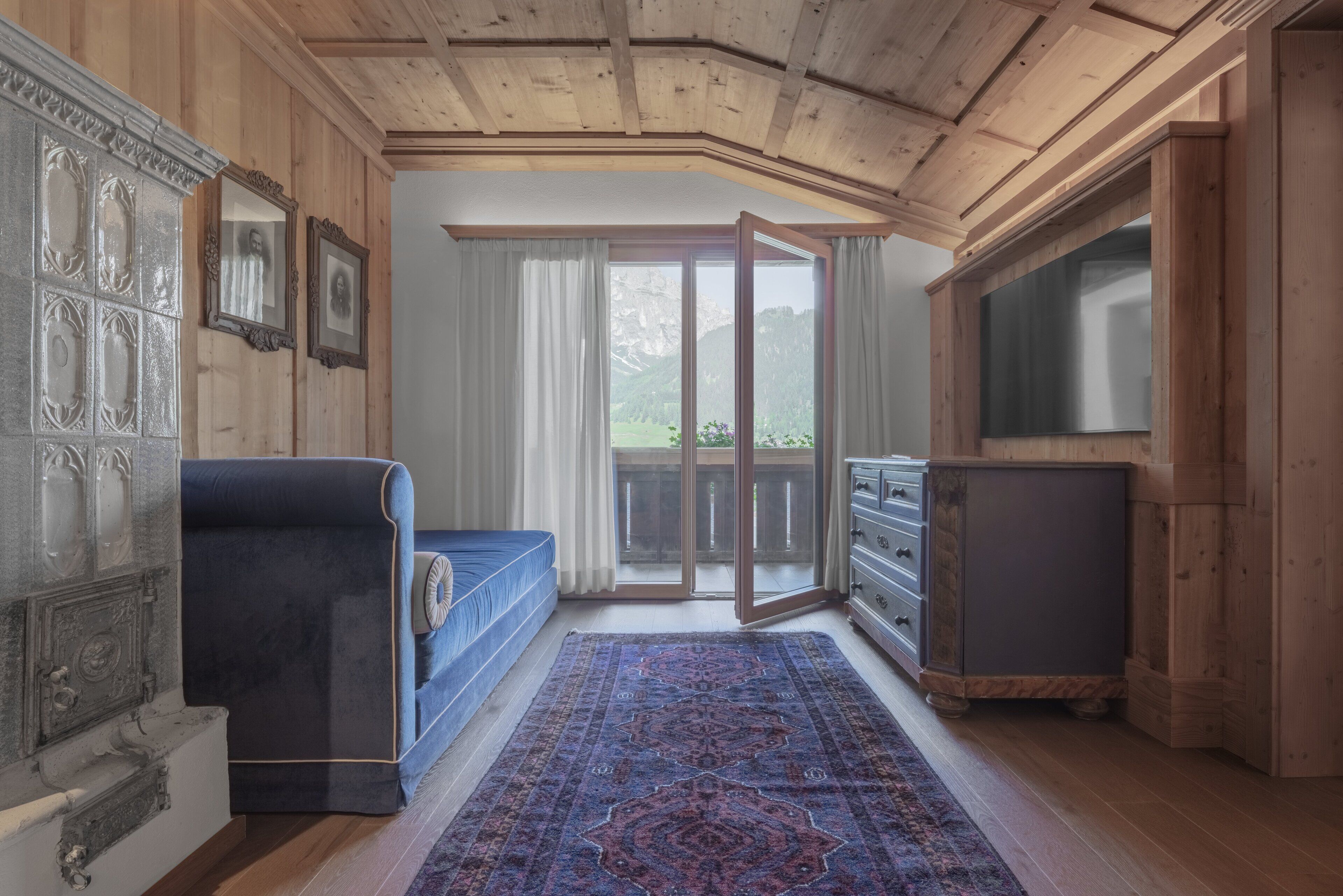 Ein Zimmer mit Holzbalkendecke, einem blauen Sofa, einem gemusterten Teppich, einer Kommode und einem großen Fenster mit Bergblick.